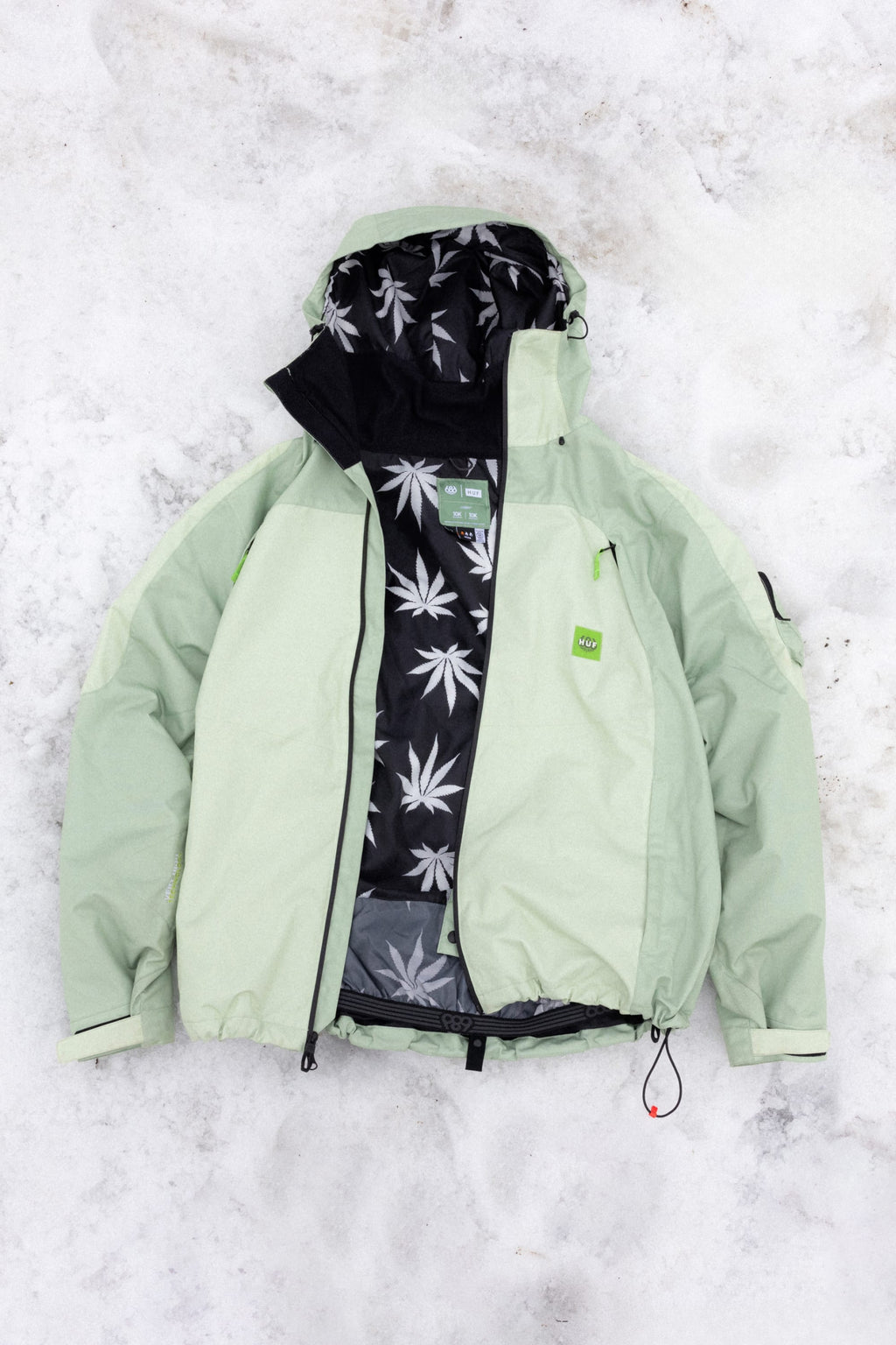 The 686 Mens HUF Double H Shell Jacket by 686 is a light green polyester waterproof jacket with black zipper details, a green logo chest tag, and a black inner lining featuring a white leaf pattern—perfect for stylish protection in any weather.