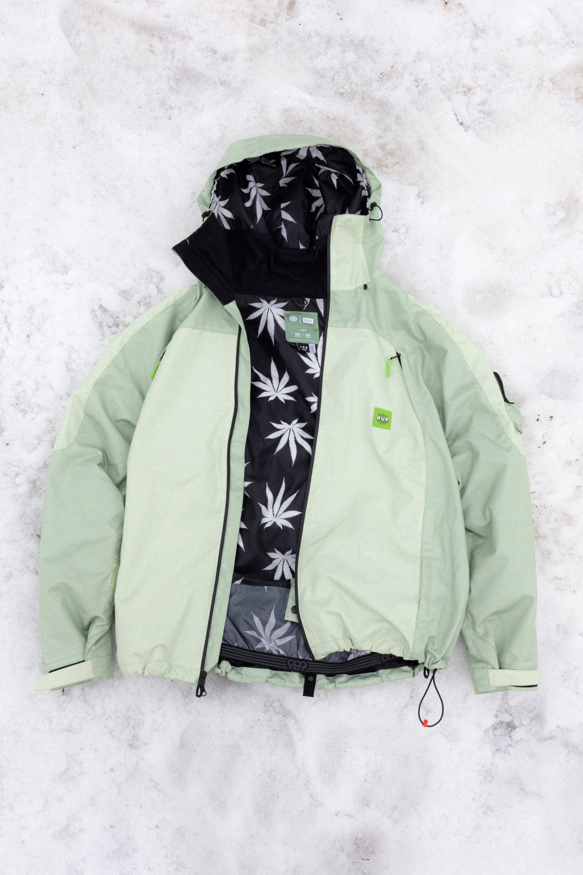 The 686 Mens HUF Double H Shell Jacket by 686 is a light green polyester waterproof jacket with black zipper details, a green logo chest tag, and a black inner lining featuring a white leaf pattern—perfect for stylish protection in any weather.