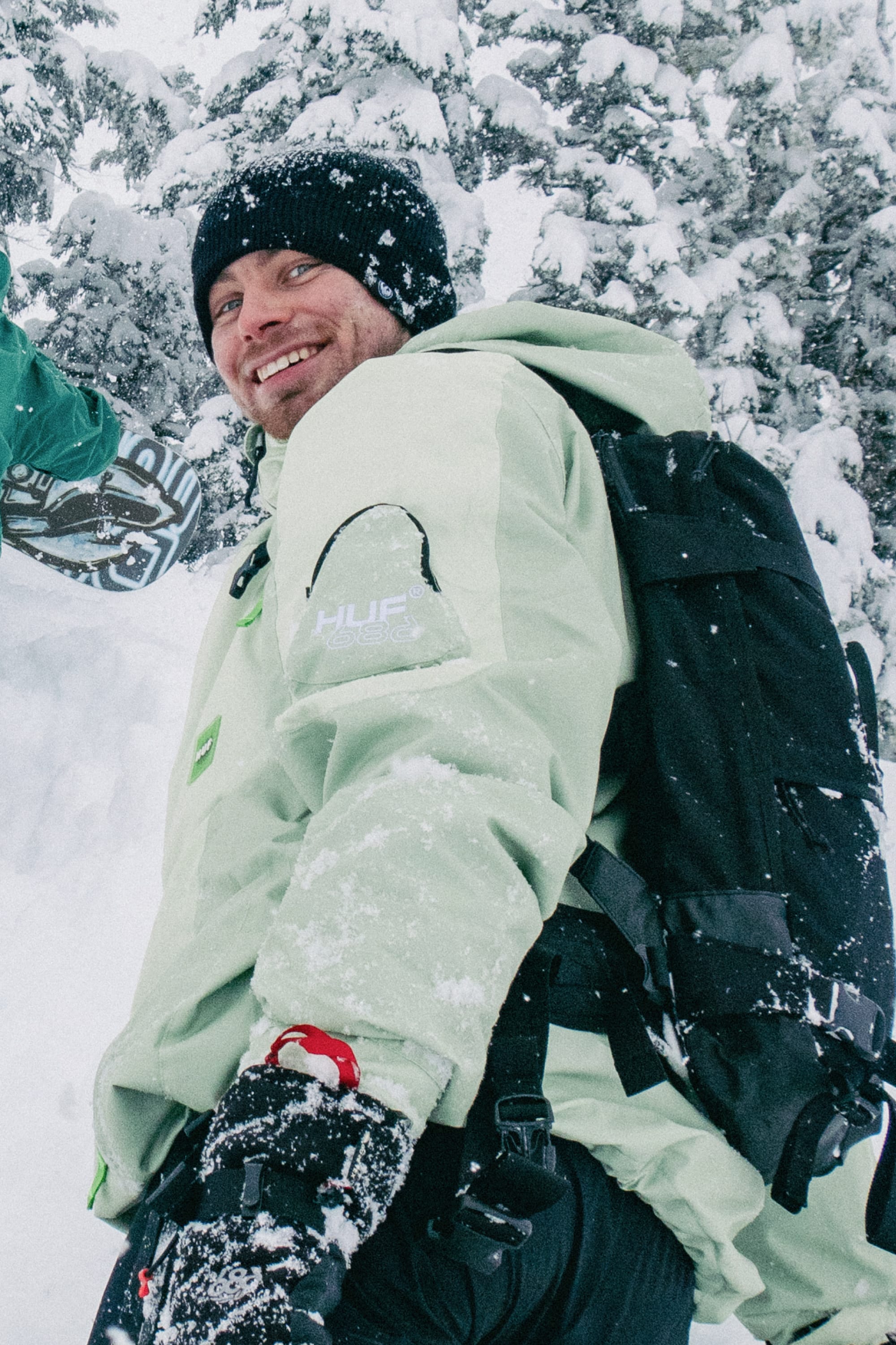 A person in a 686 Mens HUF Double H Shell Jacket, black beanie, and black backpack smiles while standing outdoors among snow-covered trees.