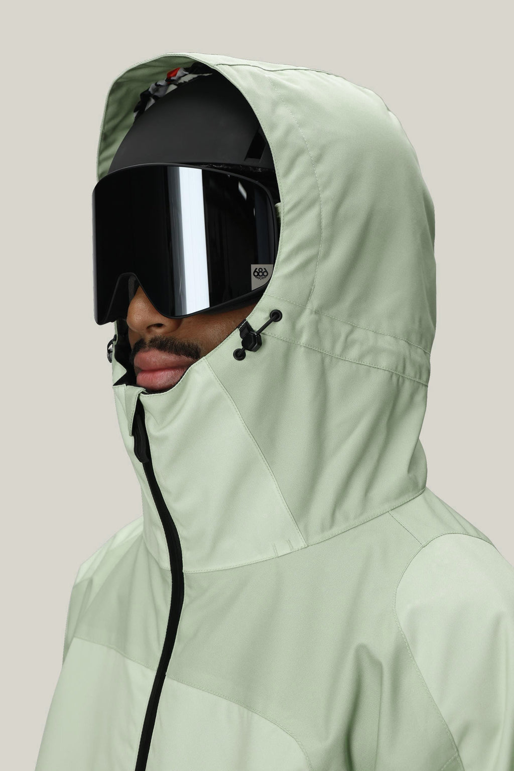 A man in a 686 Mens HUF Double H Shell Jacket (light green, waterproof, hooded) with black ski goggles and a black helmet faces left against a plain light background.