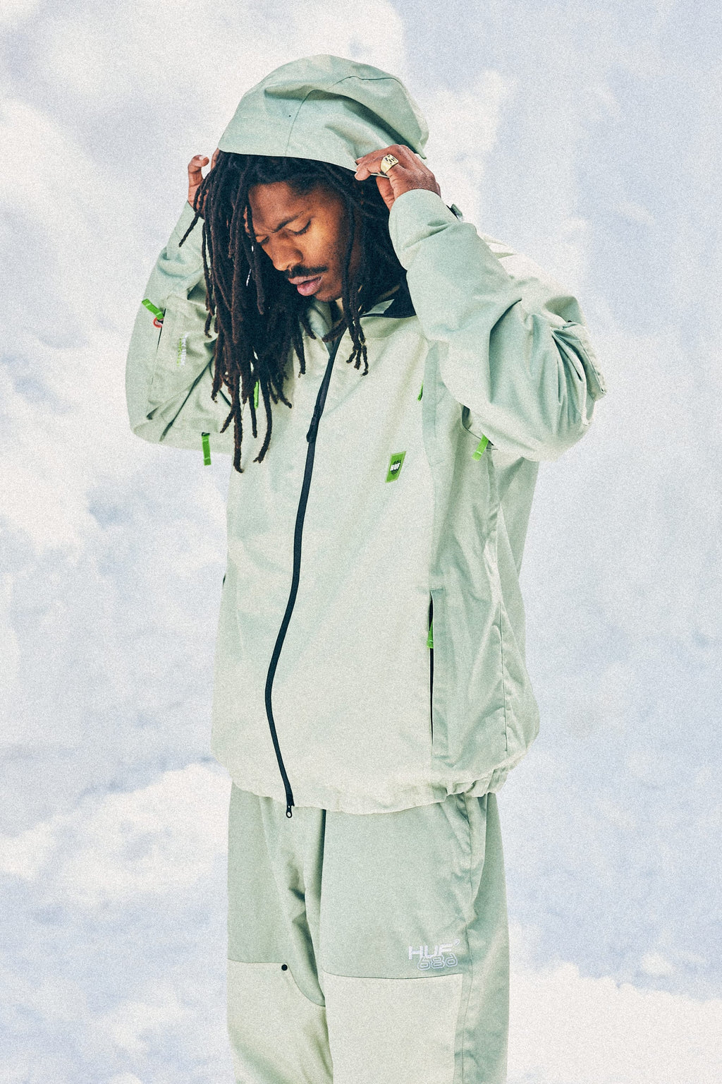 A man with long dreadlocks adjusts the hood of his 686 Mens HUF Double H Shell Jacket in light green, standing against a cloudy light blue sky. He is also wearing matching light green pants.
