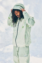 A man with long dreadlocks adjusts the hood of his 686 Mens HUF Double H Shell Jacket in light green, standing against a cloudy light blue sky. He is also wearing matching light green pants.