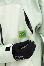 A person in a light green 686 Mens HUF Double H Shell Jacket pulls on a zipper, wearing black gloves. The chest shows the 686 logo patch on the polyester waterproof jacket.