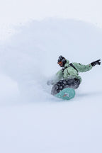 Wearing the 686 Mens HUF Double H Shell Jacket, a snowboarder carves through deep powder, sending a spray of snow behind him on a snowy slope.