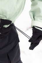 Wearing black gloves and a mint green 686 Mens HUF Double H Shell Jacket, a person tightens the adjustable hem drawstring against a snowy backdrop.