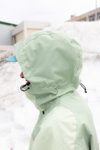 A person wearing the 686 Mens HUF Double H Shell Jacket in light green and a blue cap stands outside in a snowy area with buildings behind them, their face mostly hidden by the jackets hood.