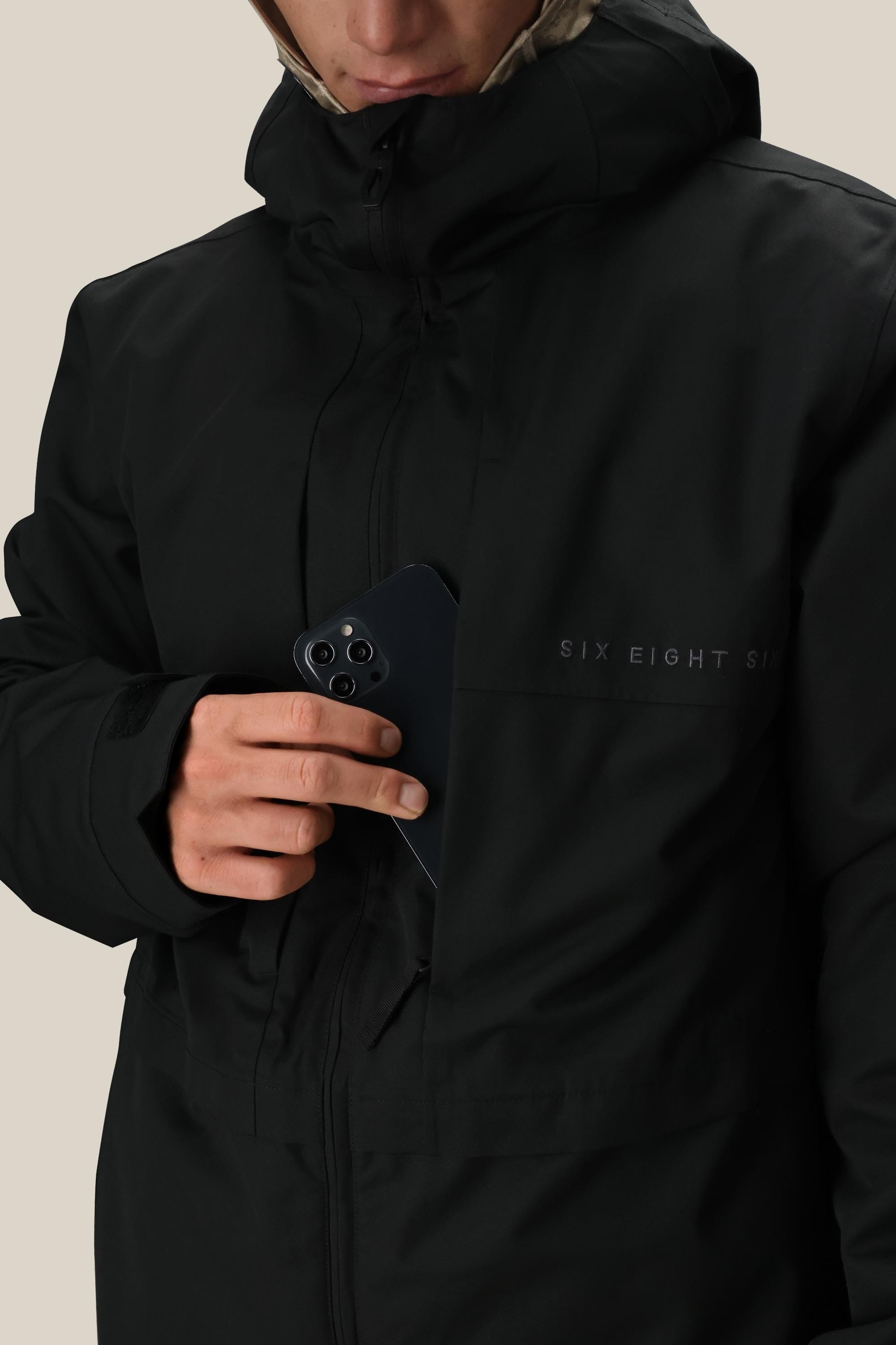 A person wearing the 686 Men's SMARTY® 3-In-1 Form Jacket in black is slipping a black smartphone into its zippered chest pocket. The jacket features the 686 logo on the chest, and the person's face is partially visible.