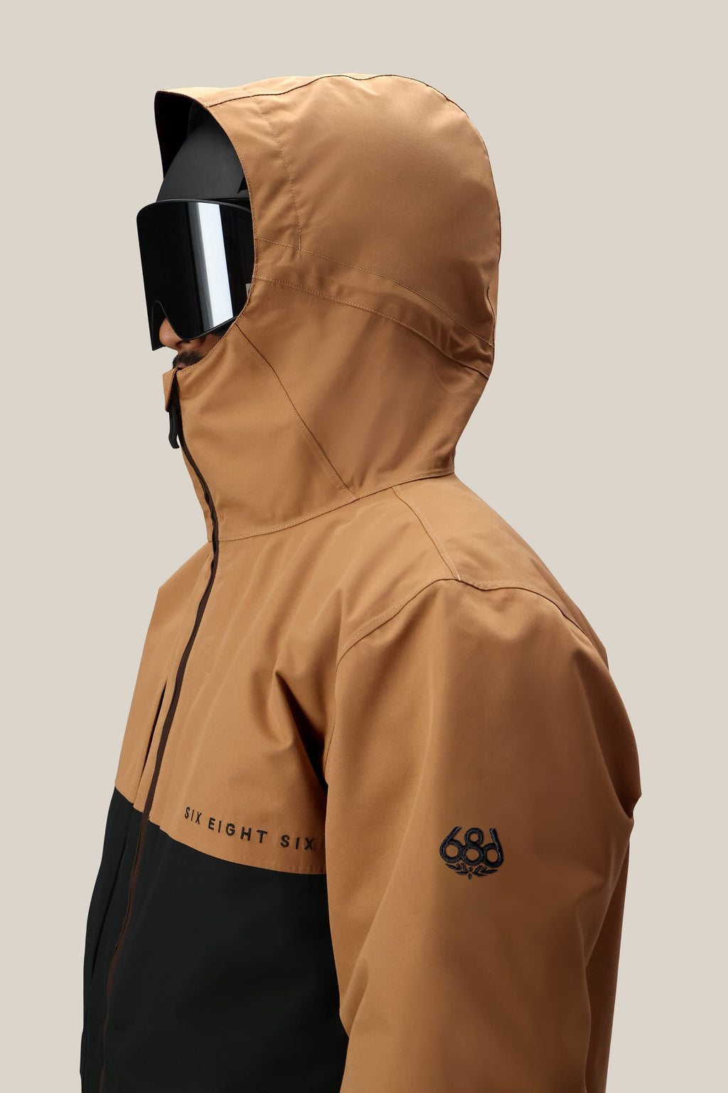 A man, shown from the side against a neutral background, wears the 686 Men's SMARTY® 3-In-1 Form Jacket in tan and black with SIX EIGHT SIX chest text, 686 sleeve logo, and large black ski goggles.