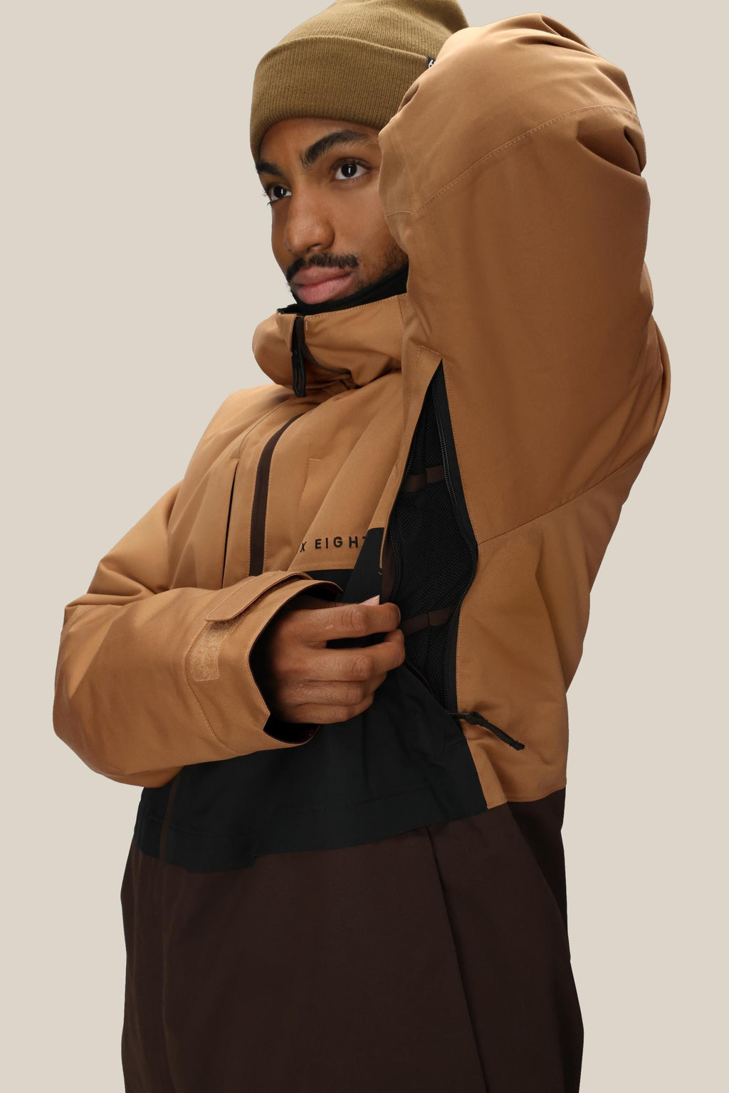 A person wearing a tan beanie and the 686 Men's SMARTY® 3-In-1 Form Jacket in brown/tan unzips an underarm vent to reveal a black mesh lining. The background is plain and light-colored.