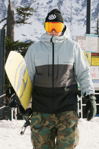 A snowboarder in a black beanie, orange goggles, and camouflage pants holds a yellow snowboard on the snow. He wears the 686 Men's SMARTY® 3-In-1 Form Jacket, with mountains and safety signs seen in the background.