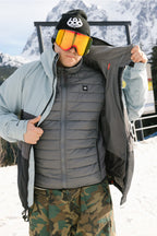 A man wearing a black beanie, orange ski goggles, camouflage pants, and the 686 Men's SMARTY® 3-In-1 Form Jacket by 686 stands on snow with mountains behind him, unzipping his jacket.