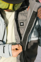 A man in ski goggles unzips his light blue-grey 686 Men's SMARTY® 3-In-1 Form Jacket, revealing the inner lining with the 686 logo and SIX EIGHT text on the chest.