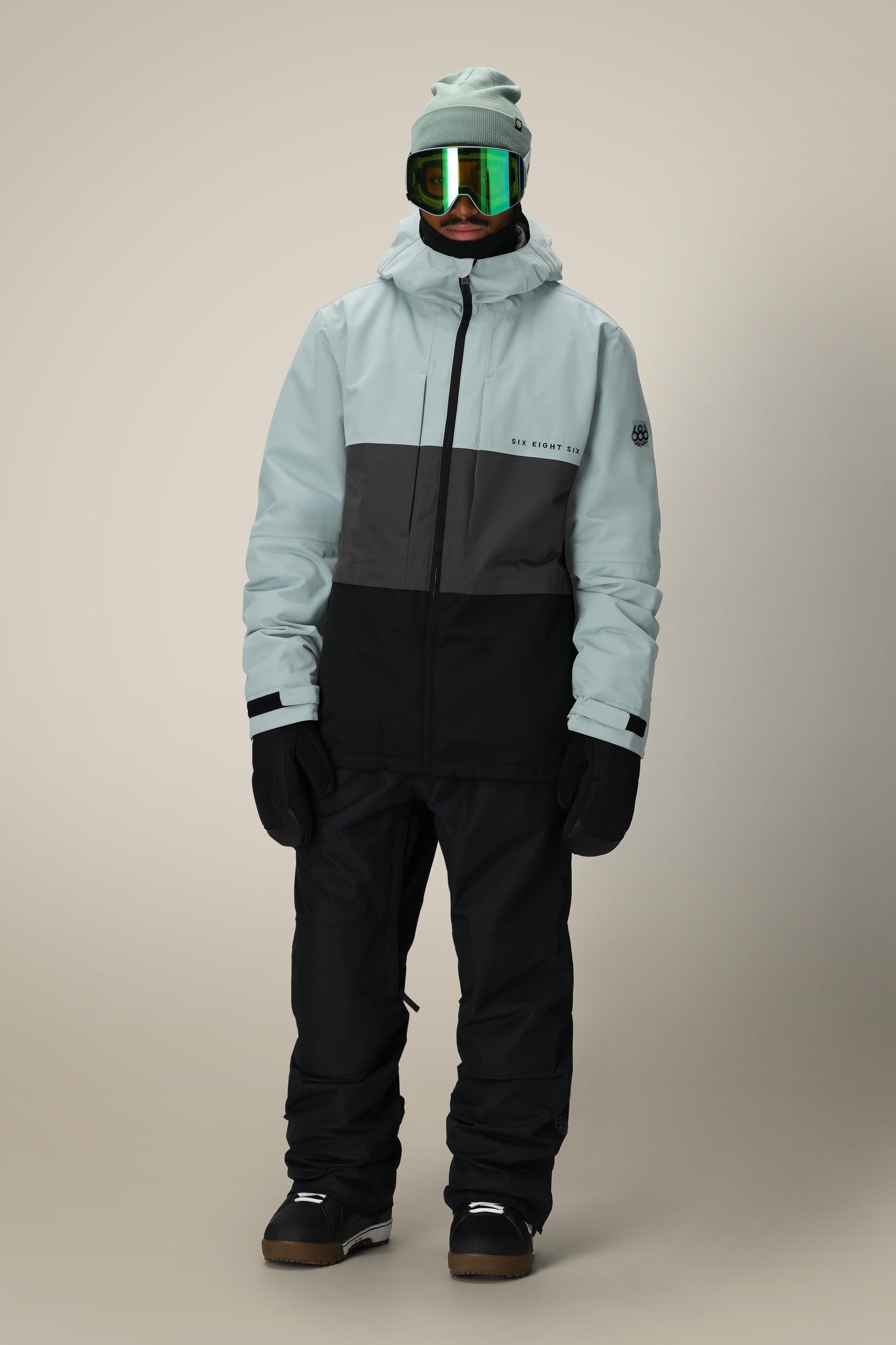 A person wearing the 686 Men's SMARTY® 3-In-1 Form Jacket by 686 in light gray and black, paired with black snow pants, gloves, a beanie, goggles with green reflective lenses, and black snow boots stands against a neutral background.