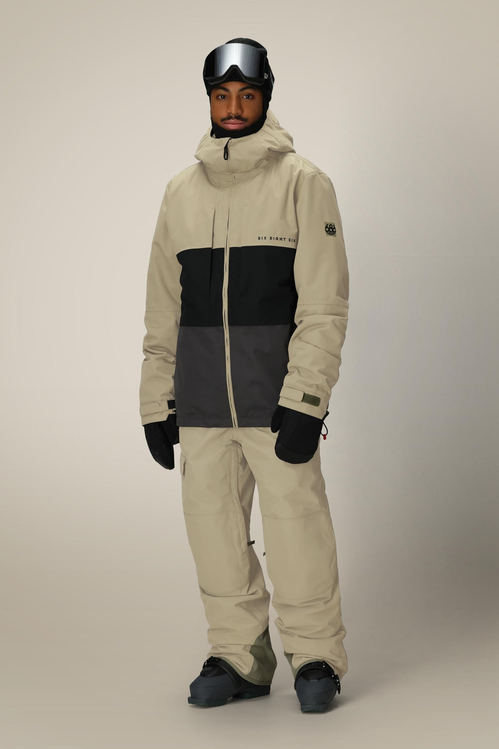 A person wearing the 686 Men's SMARTY® 3-In-1 Form Jacket in beige and black, along with ski gear such as snow pants, gloves, boots, and ski goggles, stands against a plain light background.