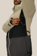 A person wearing the 686 Men's SMARTY® 3-In-1 Form Jacket in beige and black unzips an underarm vent, showing the breathable mesh lining. The two-tone jacket by 686 has a high collar, and the person's face is partially visible.