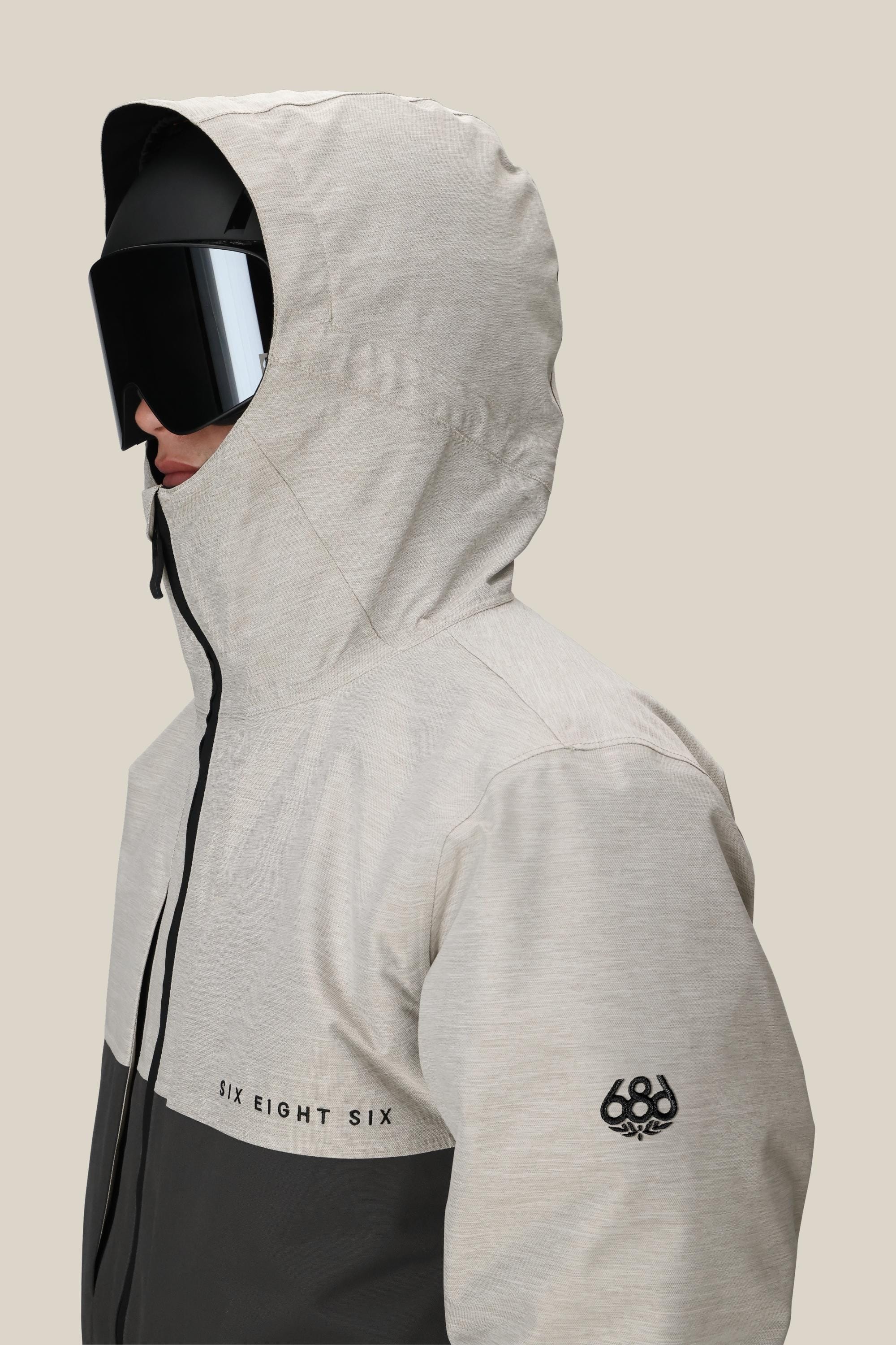 A person faces left against a plain beige background, wearing the 686 Men's SMARTY® 3-In-1 Form Jacket in light gray with black accents, black ski goggles, and the 686 logo on the sleeve.