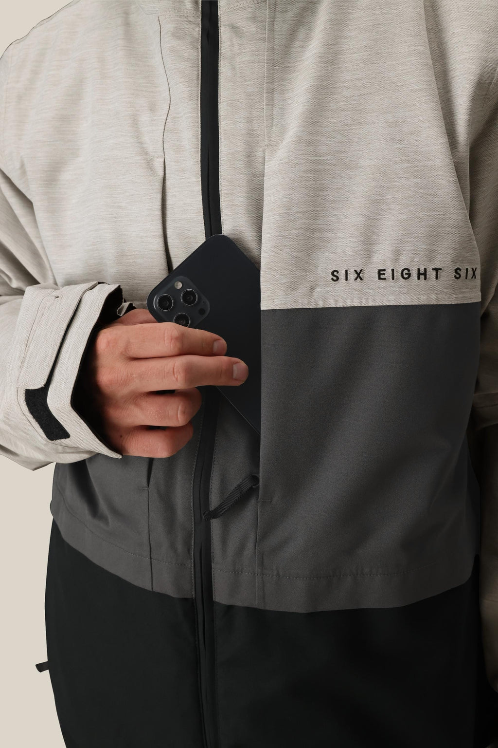 A person wearing the 686 Men's SMARTY® 3-In-1 Form Jacket in grey and black puts a black smartphone into the chest pocket.
