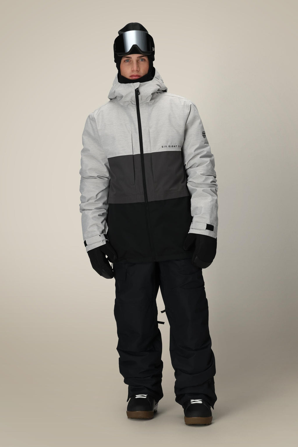 A man stands facing forward against a plain beige background, wearing the 686 Men's SMARTY® 3-In-1 Form Jacket in black/gray, along with black snow pants, black gloves, ski goggles, and a black beanie.
