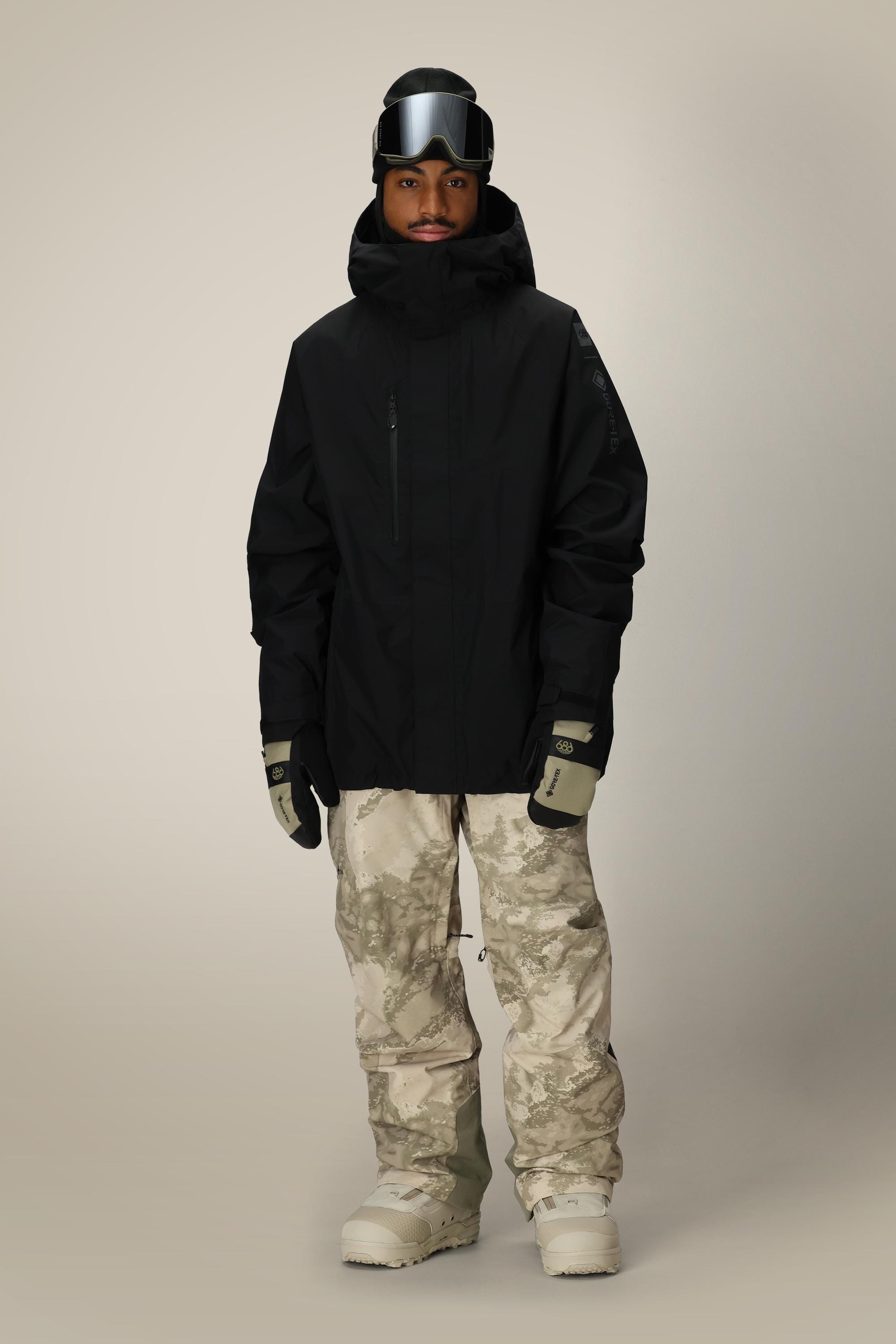 A person wearing the 686 Mens GORE-TEX Core Shell Jacket, beige and gray camo snow pants, beige gloves, white snow boots, and ski goggles stands facing forward against a plain light background.