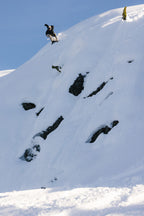 A snowboarder wearing the 686 Mens GORE-TEX Core Shell Jacket rides down a steep, snow-covered mountain slope with exposed rocks under a clear blue sky.