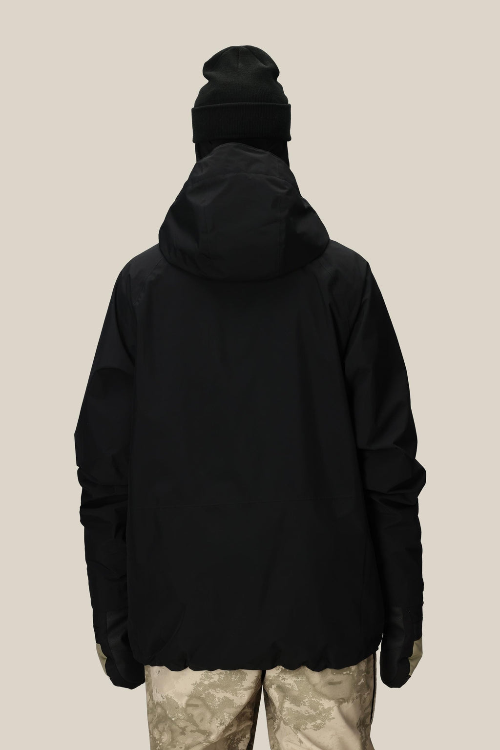 A person wearing the 686 Mens GORE-TEX Core Shell Jacket with a hood, a black beanie, and patterned beige pants stands with their back to the camera against a plain light background.