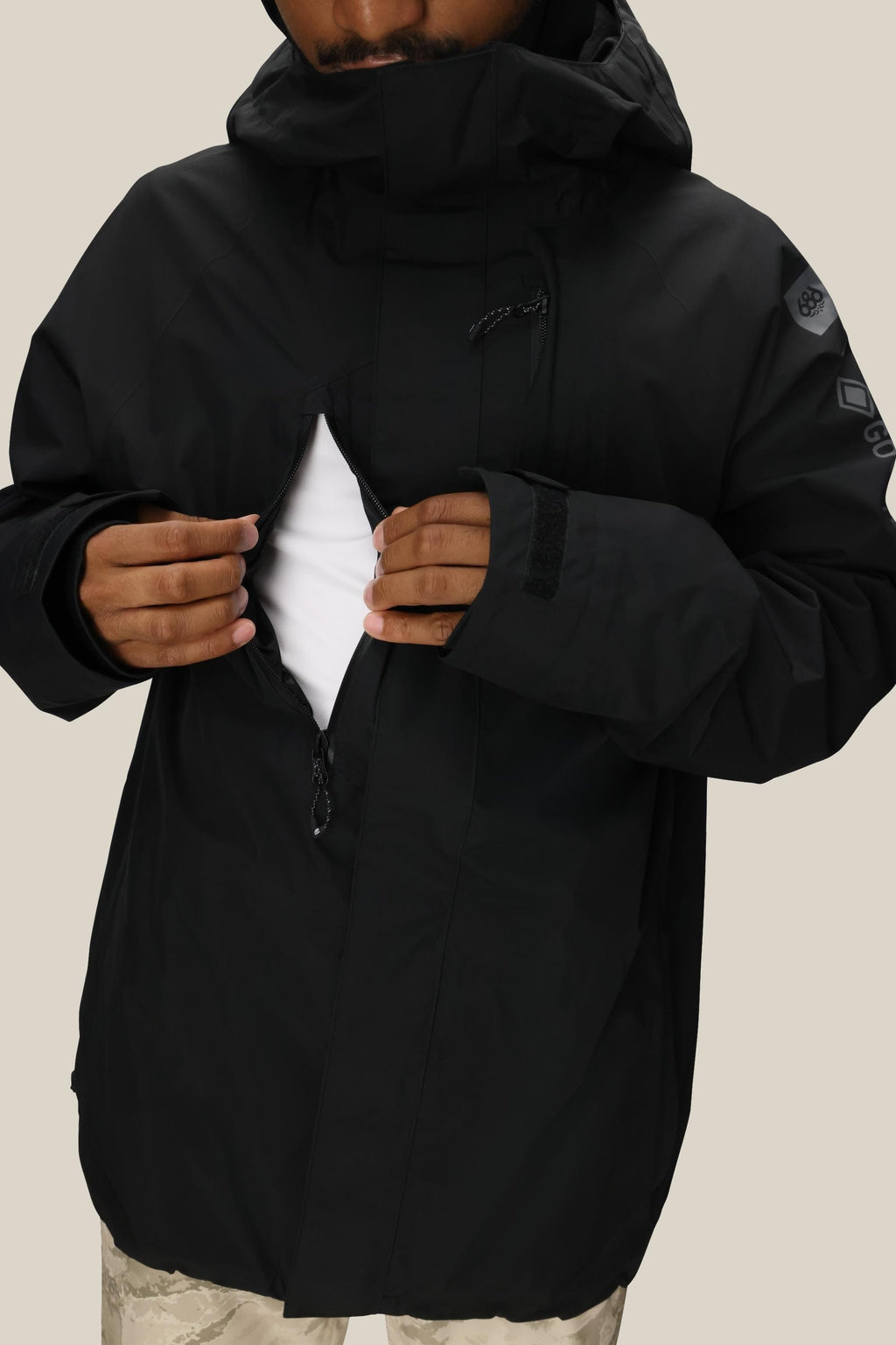 A person in the 686 Mens GORE-TEX Core Shell Jacket opens a zippered chest pocket, revealing a white inner layer. Only the lower face is visible against a plain, light background.