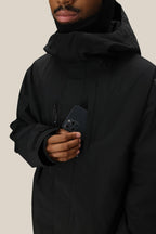 A man wearing a 686 Mens GORE-TEX Core Shell Jacket in black slips a black smartphone into its chest pocket. The background is plain beige, and only the lower half of his face is shown.