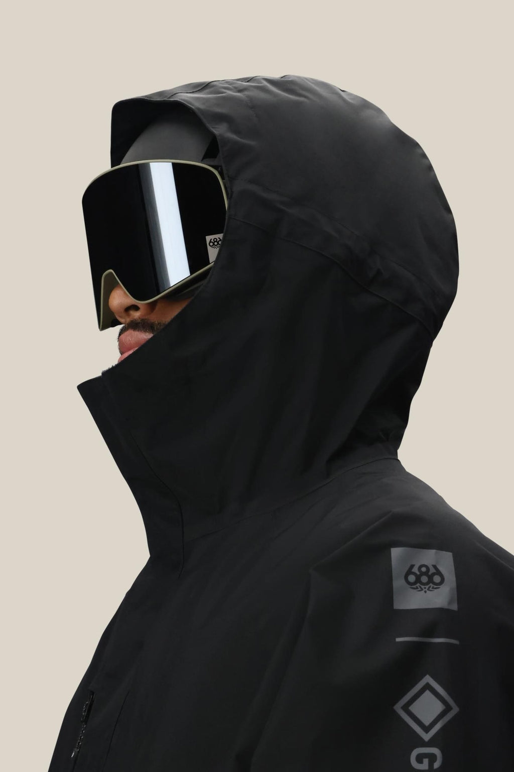 A man in a 686 Mens GORE-TEX Core Shell Jacket with sleeve logos, large black ski goggles, and a helmet is shown in side view against a plain beige background.