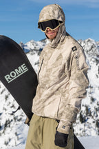 A snowboarder in a 686 Mens GORE-TEX Core Shell Jacket, tan camo gear, black gloves, and goggles holds a black ROME snowboard with snowy mountains in the background.