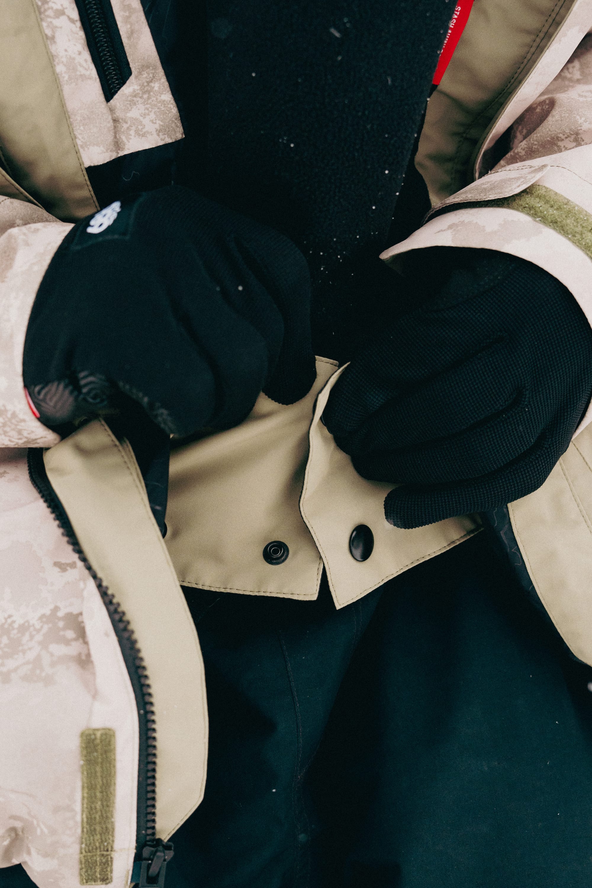 A person wearing black gloves fastens a snap button on the 686 Mens GORE-TEX Core Shell Jacket. Only their hands and part of the 686 jacket are visible.