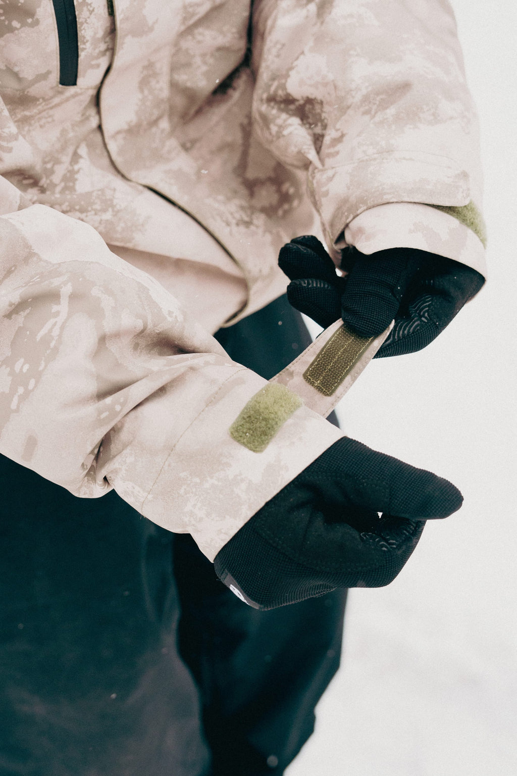 A person wearing black gloves adjusts the Velcro strap on the sleeve of a 686 Mens GORE-TEX Core Shell Jacket in beige camo, with snow visible in the background.