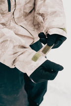 A person wearing black gloves adjusts the Velcro strap on the sleeve of a 686 Mens GORE-TEX Core Shell Jacket in beige camo, with snow visible in the background.