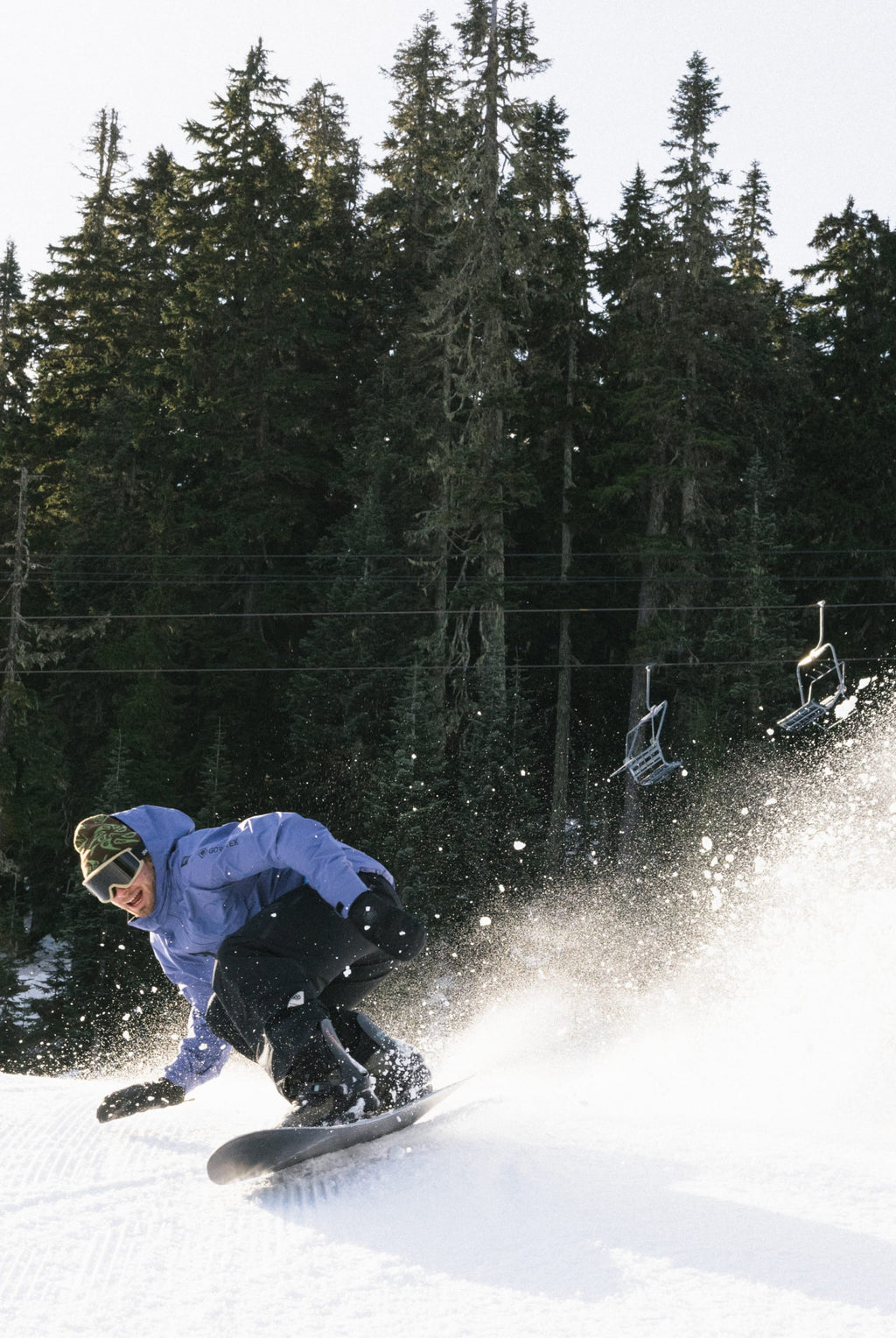 A snowboarder in a 686 Mens GORE-TEX Core Shell Jacket and black pants carves down a snowy slope, kicking up powder with tall evergreens and empty ski lift chairs in the background.