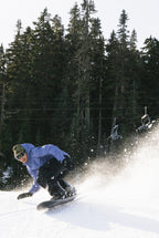 A snowboarder in a 686 Mens GORE-TEX Core Shell Jacket and black pants carves down a snowy slope, kicking up powder with tall evergreens and empty ski lift chairs in the background.