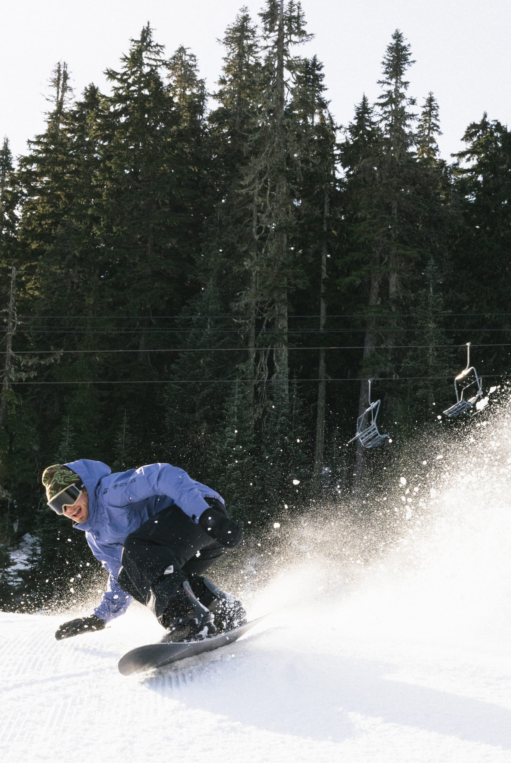 A snowboarder in a 686 Mens GORE-TEX Core Shell Jacket and black pants carves down a snowy slope, kicking up powder with tall evergreens and empty ski lift chairs in the background.