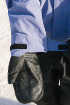 A person wearing a blue 686 Mens GORE-TEX Core Shell Jacket stands on snow, extending their gloved hand toward the camera.