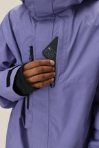 A man wearing the 686 Mens GORE-TEX Core Shell Jacket in purple puts a black smartphone with three camera lenses into the jackets zippered chest pocket.