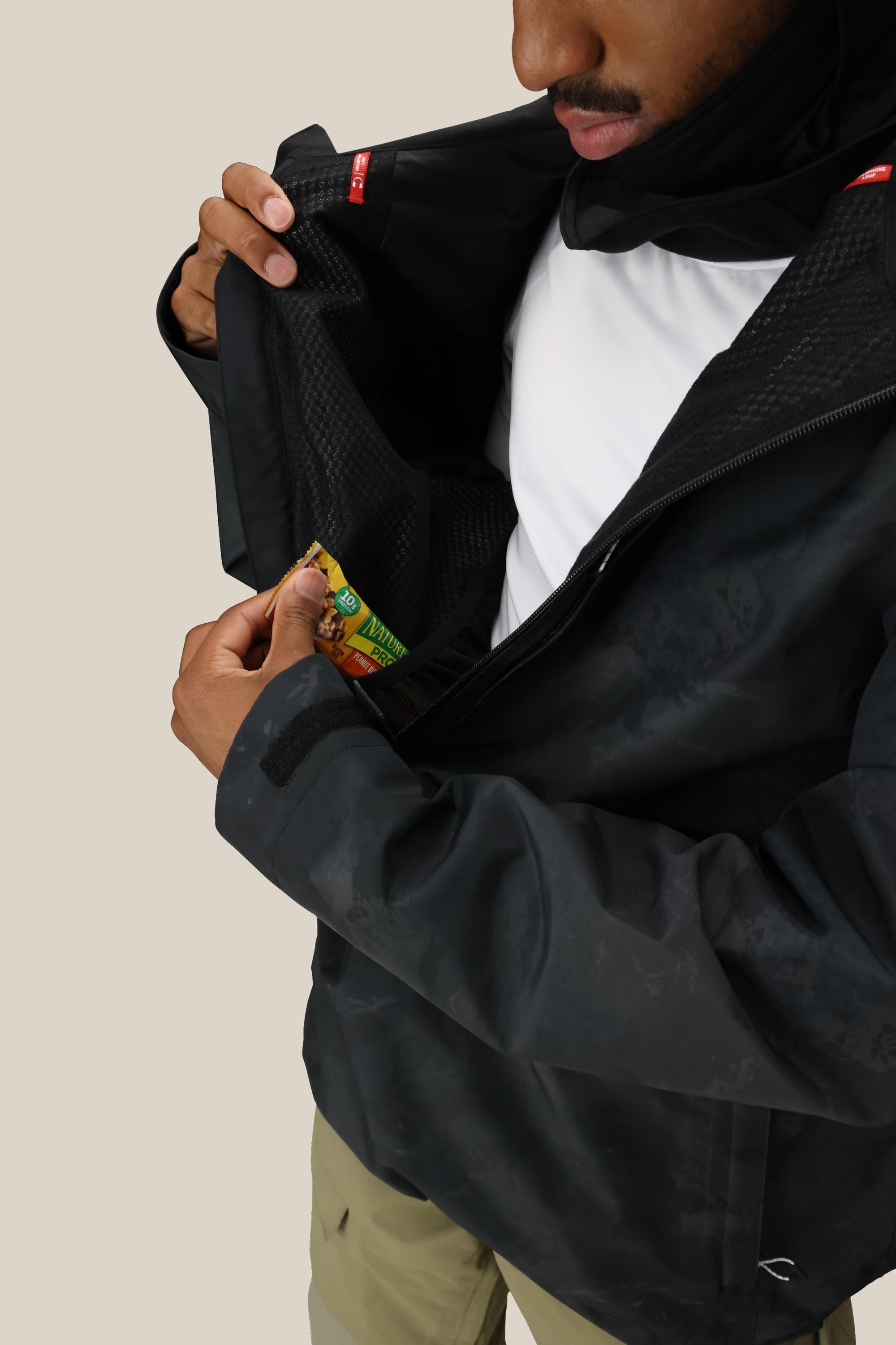 A person in the 686 Mens GORE-TEX Fragment Shell Anorak (black) with a white shirt places a granola bar into the inner chest pocket. Their face is partially visible against a plain light beige background.