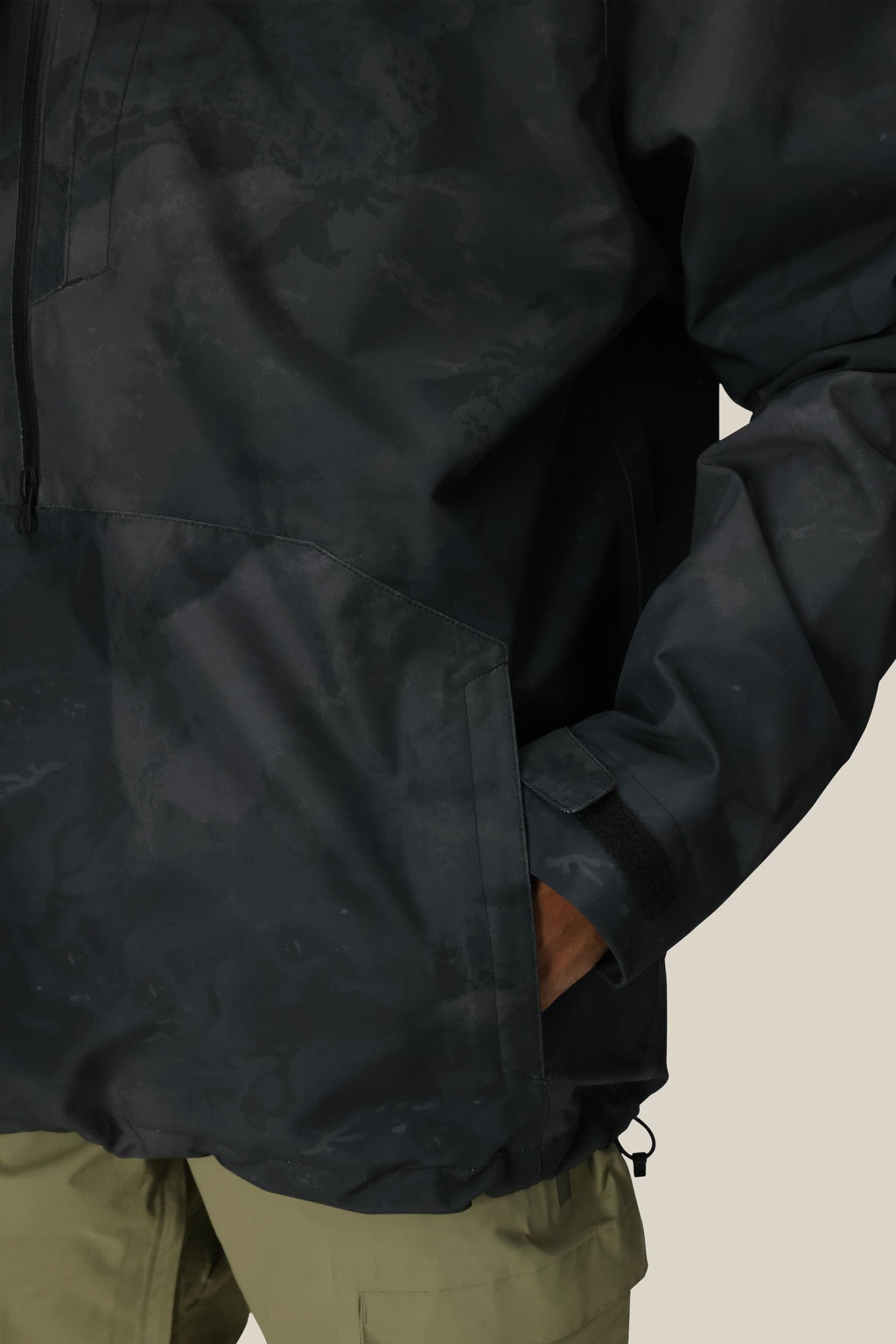 A person wears the 686 Mens GORE-TEX Fragment Shell Anorak with a dark pattern, right hand in the pocket. Only part of the jacket and olive green pants are visible against a neutral background.