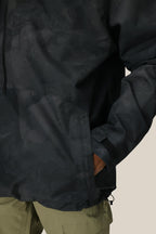 A person wears the 686 Mens GORE-TEX Fragment Shell Anorak with a dark pattern, right hand in the pocket. Only part of the jacket and olive green pants are visible against a neutral background.