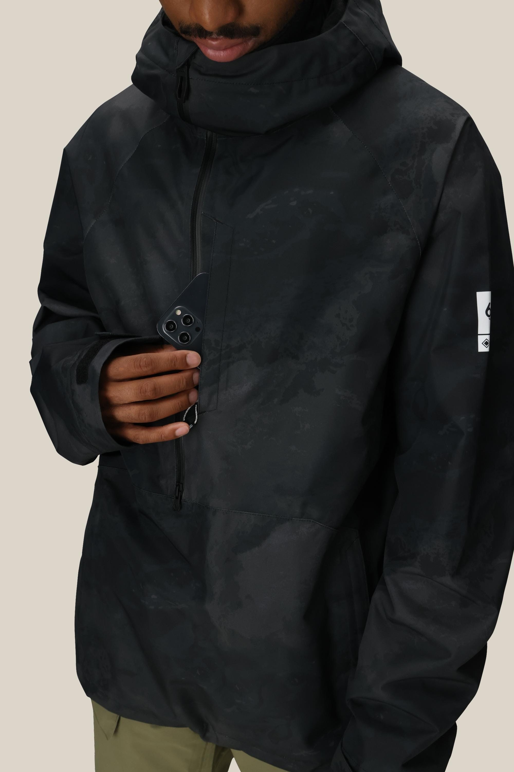 A person wearing the 686 Mens GORE-TEX Fragment Shell Anorak in black, featuring a subtle camouflage pattern and a white sleeve logo, holds a smartphone in the chest pocket and pairs it with olive green pants.