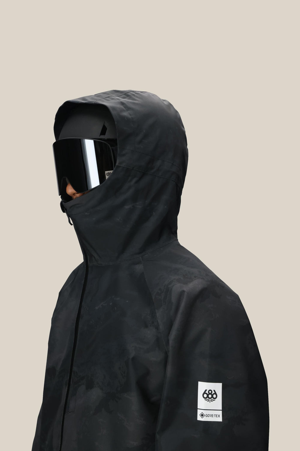 A man wearing the 686 Mens GORE-TEX Fragment Shell Anorak in black with a white logo patch on the sleeve and large black ski goggles, shown from the side against a plain beige background.