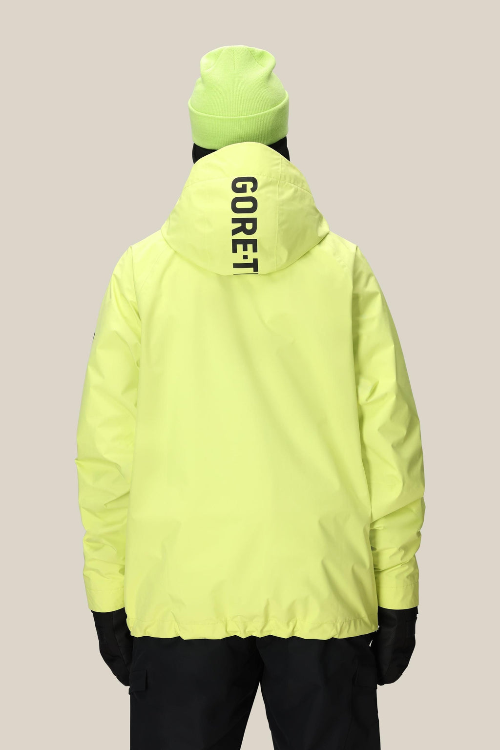A person stands facing away against a plain beige background, wearing the 686 Mens GORE-TEX Fragment Shell Anorak in bright yellow with GORE-TEX on the hood and a matching yellow beanie.