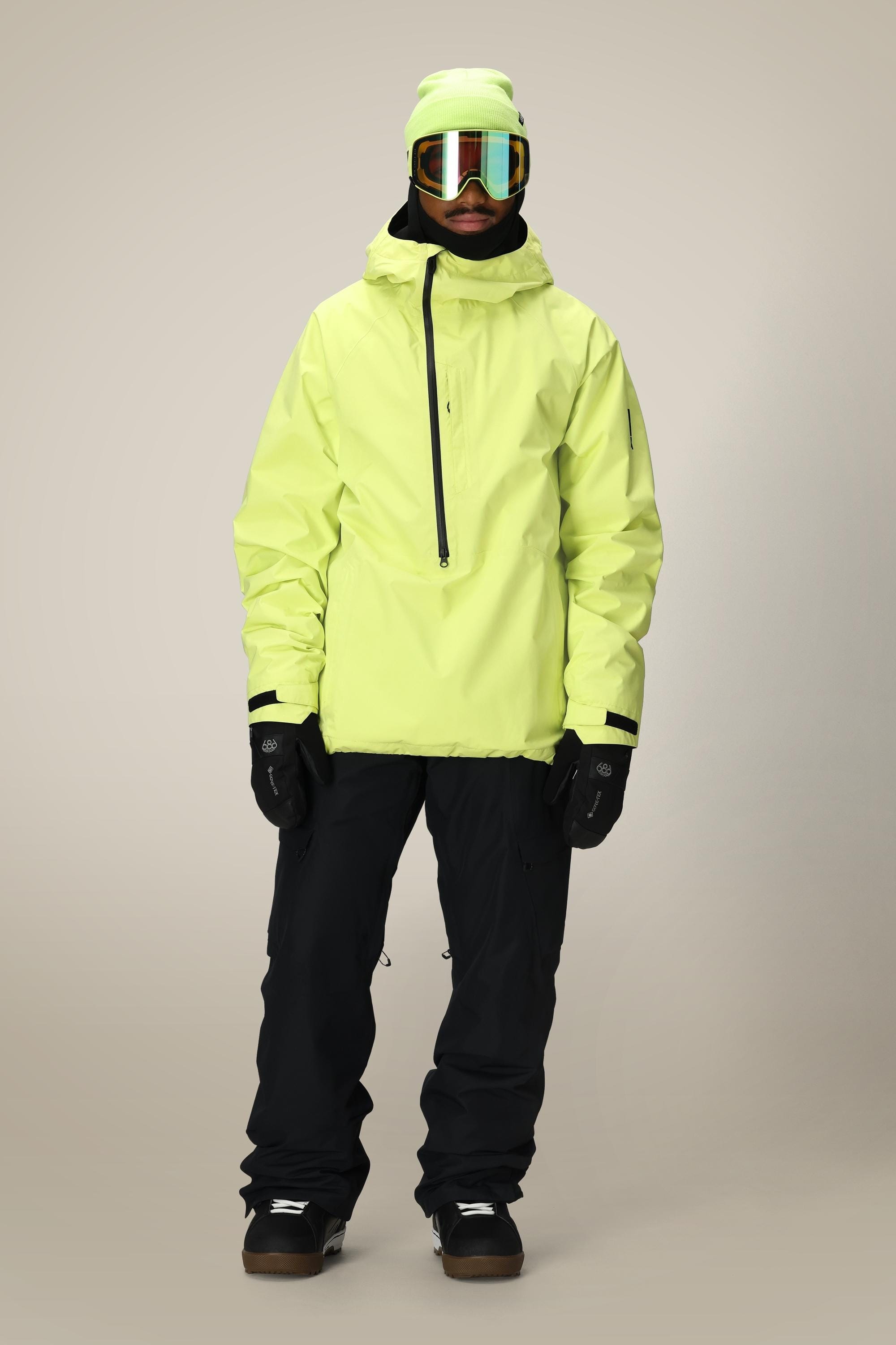 A person stands facing forward wearing the 686 Mens GORE-TEX Fragment Shell Anorak in light yellow, black snow pants, black gloves and boots, a yellow-green beanie, and reflective ski goggles against a plain background.