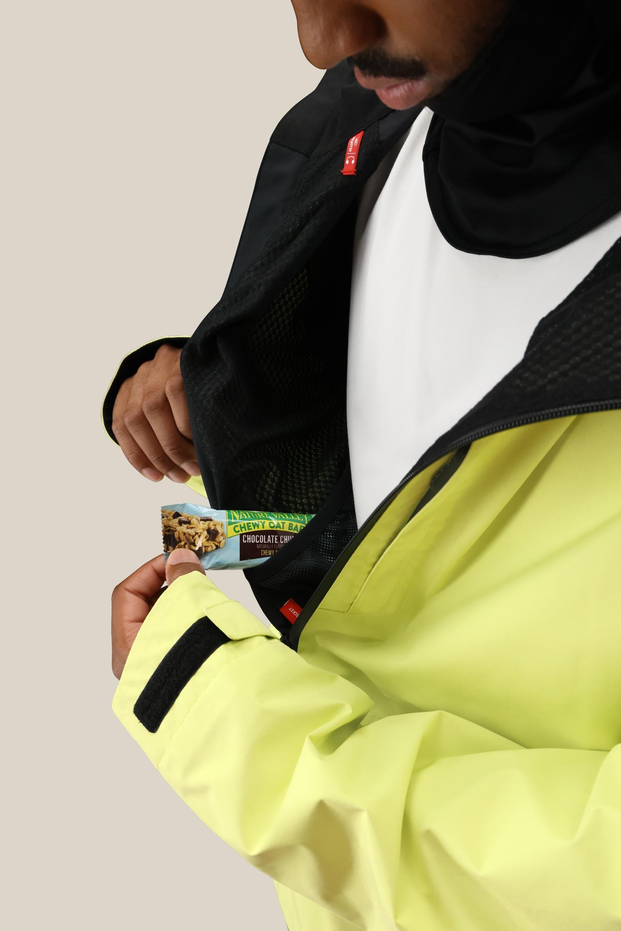 A person wearing the 686 Mens GORE-TEX Fragment Shell Anorak in yellow and black puts a chocolate chip granola bar into their inner pocket. The cropped image shows only the lower face and torso.