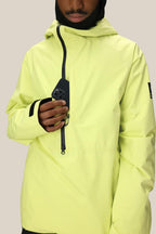 A person wearing the 686 Mens GORE-TEX Fragment Shell Anorak in bright yellow partially unzips the chest pocket to place or remove a black smartphone. The individual’s face is partly visible, and the background is plain light beige.