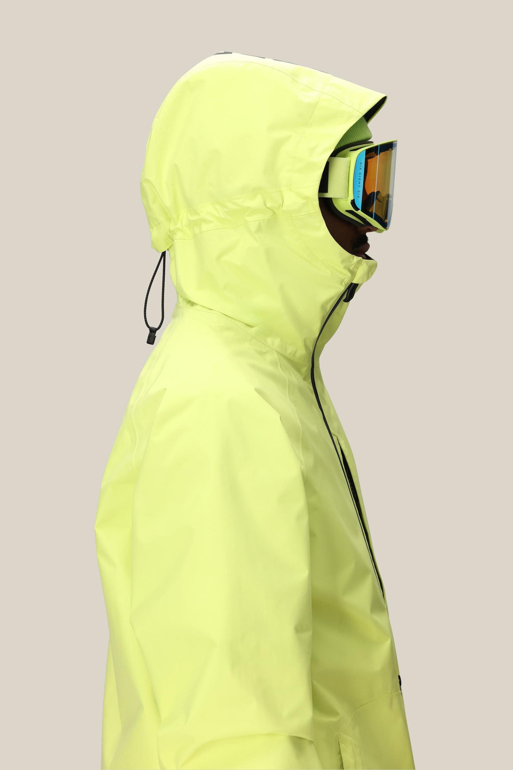 Person in profile wearing the 686 Mens GORE-TEX Fragment Shell Anorak in bright yellow with hood up and drawstring cinched, plus reflective ski goggles, against a plain beige background.