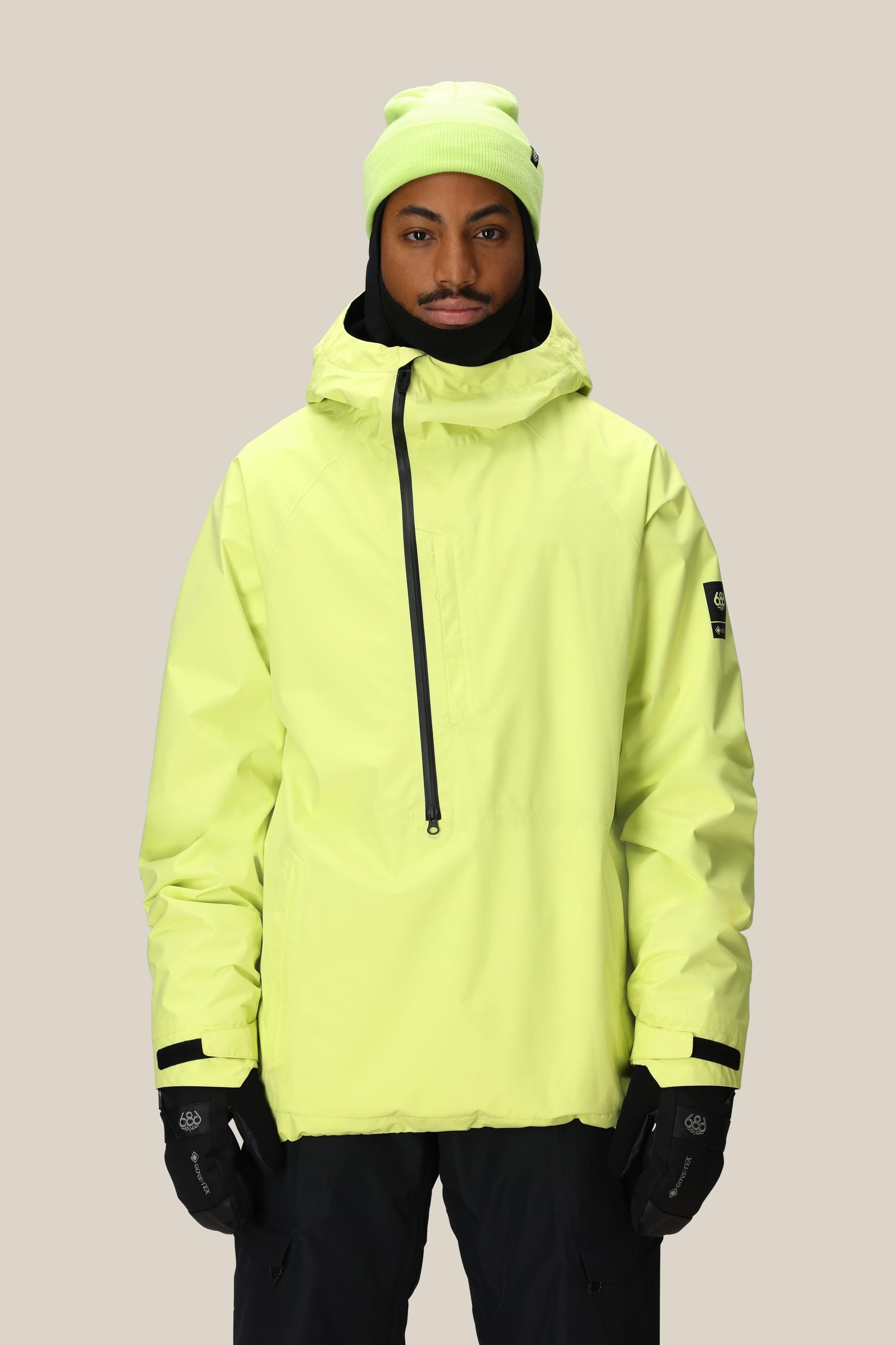 A man in a neon yellow 686 Mens GORE-TEX Fragment Shell Anorak with hood, matching beanie, black gloves, and black pants stands against a light gray background.
