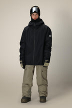 A person wearing the 686 Mens Gateway Shell Jacket, beige snow pants, gloves, dark boots, a helmet, and ski goggles stands against a plain light background.