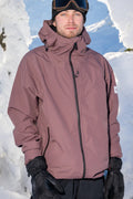 A person wearing the 686 Mens Gateway Shell Jacket by 686, along with black gloves and ski goggles, stands outdoors surrounded by snow and bright sunlight.